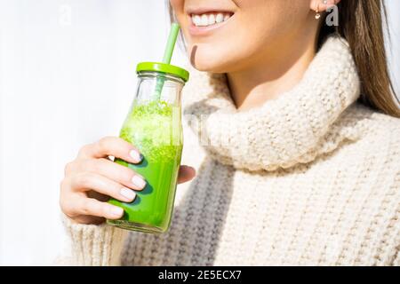 Donna sorridente in pullover di lana che beve frullato disintossicazione o succo di frutta fatti in casa verde. Mangiare sano Foto Stock