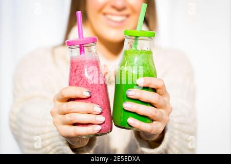 Donna sorridente felice in pullover di lana che dà 2 bottiglie di frullato fresco rosa e verde Foto Stock