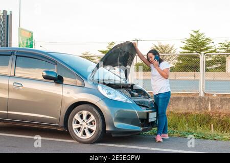 Ragazze asiatiche che tengono un telefono cellulare camminando intorno all'automobile, umore stressante durante le ore serali. Lungo l'autostrada perché la sua auto ha rotto fare Foto Stock