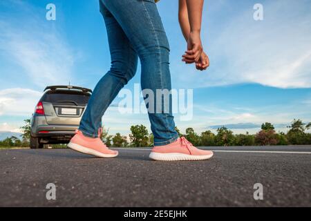 Donne che indossano jeans sneakers binding sulla strada, auto parcheggiata lungo la strada di giorno, il cielo blu luminoso e sfondo nuvole Foto Stock