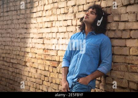 UN GIOVANE CON I CAPELLI RICCI APPOGGIATI SULLA PARETE E. ASCOLTO DI MUSICA Foto Stock