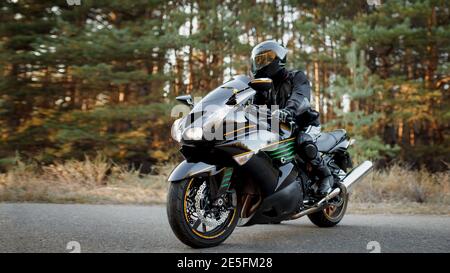 Conducente di motocicletta in una tuta in pelle e seduto su un moto da solo su una strada asfaltata su uno sfondo forestale Foto Stock