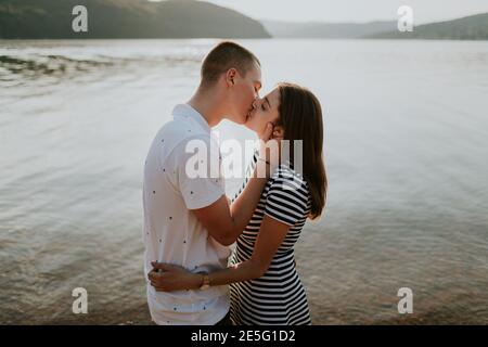 Primo piano di coppia in amore baciando al lago al tramonto. Ritratto di fidanzata e fidanzata che abbracciano e baciano sulla spiaggia nella calda sera d'estate Foto Stock