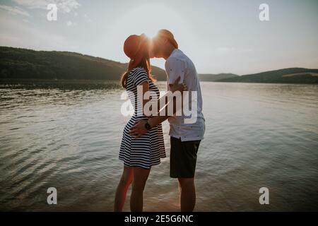 Coppia innamorata baciarsi sulla spiaggia al tramonto. Ritratto di giovane uomo e donna che abbracciano e baciano al lago contro il sole della sera.