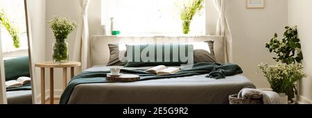 Camera da letto luminosa interna in vera foto con fiori freschi, finestra e letto matrimoniale con libro aperto, tea Cup e coperta verde Foto Stock