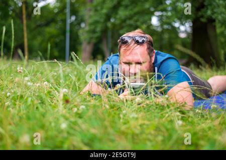 uomo adulto che tiene uno smartphone mentre si sdraiò su erba verde dentro parco cittadino Foto Stock