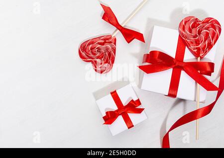 San Valentino sfondo giorno - scatole regalo bianche con arco di seta rossa, nastro, cuori di lecca su tavolo di legno bianco, spazio copia. Foto Stock