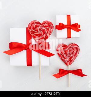 Love Holiday background - scatole regalo bianche con arco rosso, cuori di lecca dolce su tavola di legno bianco, quadrato. Foto Stock