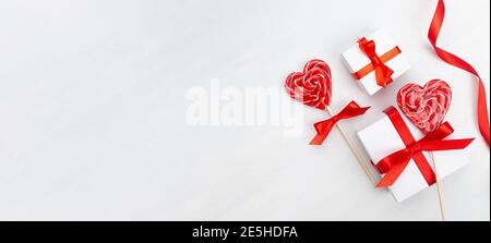 Banner giorno di San Valentino - scatole regalo bianche con arco in seta rossa, nastro, cuori di lecca su tavolo in legno bianco, spazio copia. Foto Stock