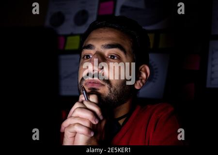 Il giovane sta pensando davanti al suo laptop a. le ore di straordinario nel suo ufficio buio Foto Stock