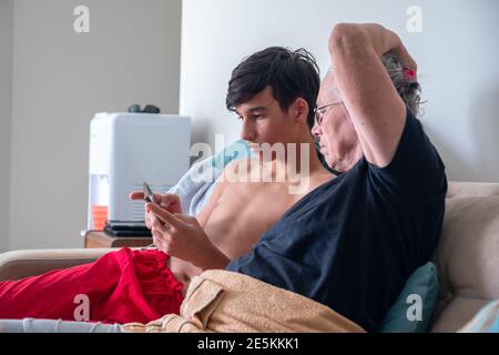 Giovane maschio che insegna sua nonna sui telefoni cellulari moderni e. nuove applicazioni in casa mentre si è seduti in un divano Foto Stock