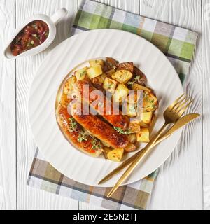 bangers e patate arrostite su un piatto bianco con sugo di cipolla in una caraffa su un tavolo di legno testurizzato bianco, piatto Foto Stock