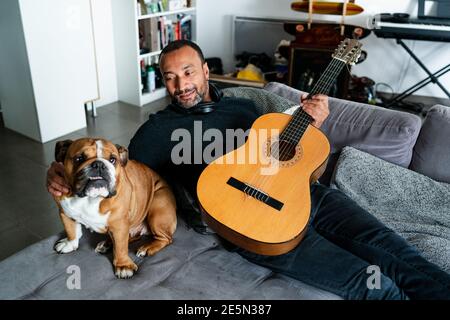 Felice uomo di mezza età con il suo amico bulldog sta giocando il chitarra a casa Foto Stock