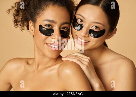 Ritratto di bellezza di due adorabili giovani donne in posa con applicato nero sotto gli occhi cerotti, in piedi insieme isolato su sfondo beige. Concetto di routine per la cura della pelle Foto Stock