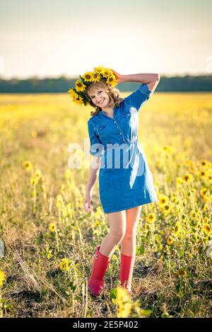 Carina ragazza sorridente dai capelli rossi nel campo pieno di girasoli, corona di girasoli. Vestita in un abito-camicia in denim e stivali di gomma rossa. Foto Stock