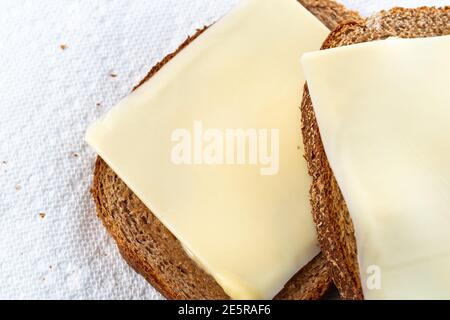 Vista ravvicinata dall'alto di due fette di pane di grano leggermente tostato con margarina e formaggio a fette su asciugamani di carta bianca con luce naturale. Foto Stock