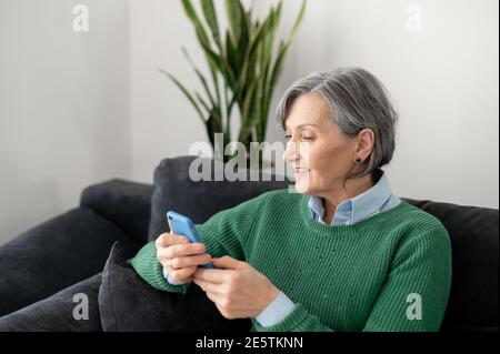 Donna anziana matura che indossa un ponticello verde, seduto sul divano nel soggiorno, tenendo un telefono cellulare e in attesa dell'applicazione Foto Stock