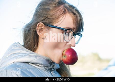 Donna caucasica che indossa occhiali tiene una mela matura rossa in bocca, raccolto, raccolta di mele, snack sano Foto Stock