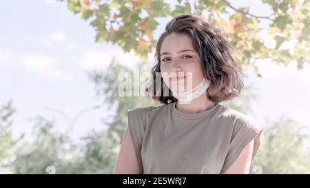 Ritratto di giovane donna di charme in maschera medica. La sua maschera facciale è sotto il mento. Nuova normalità dopo la pandemia di covid-19. Foto Stock