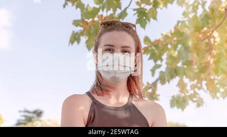 Ritratto di giovane donna di charme in maschera medica guarda la macchina fotografica, nuova normalità dopo covid-19 pandemia. Riprese all'aperto. Stili di vita sani Foto Stock