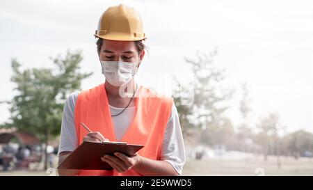 Giovane lavoratore di costruzione maschile con casco giallo e maschera facciale prendendo appunti in cantiere. Nuova normalità dopo l'epidemia di covid-19. Foto Stock