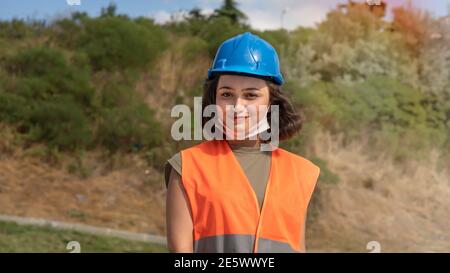 Ritratto di una giovane donna allegra ingegnere o lavoratore in un uniforme e protettivo cappello blu, sorridente e guardando la macchina fotografica in cantiere Foto Stock