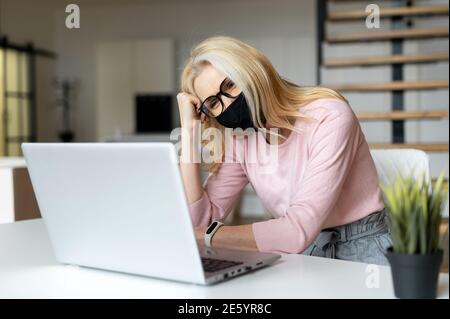 Preoccupato e stanco dipendente di ufficio femminile che indossa una maschera medica protettiva utilizzando un computer portatile indoor, una donna imprenditore è triste, la sua attività è a causa di male Foto Stock