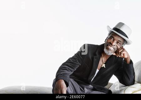 Felice l'uomo senior in fedora seduto sulla chaise longue contro uno sfondo bianco Foto Stock