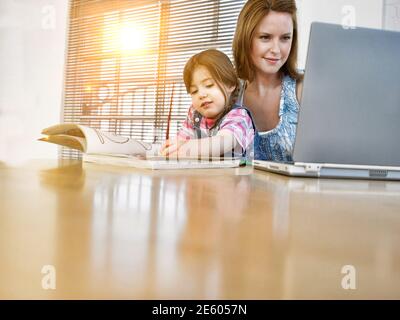 Giovane donna che lavora con il laptop con sua figlia che si colorano sopra prenota Foto Stock