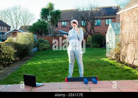 Donna che beve acqua e che parla con un addestratore durante una pausa di allenamento fitness online all'aperto. Fare fitness in giardino, cortile. Un sano Foto Stock