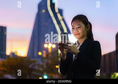 Donna di lavoro dirigente asiatica che utilizza un telefono cellulare in strada con uffici in background di notte a Bangkok, Thailandia. Foto Stock