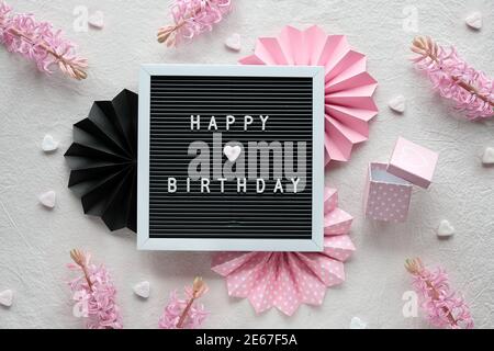 Testo buon compleanno su tabellone, carta intestata. Piatto creativo posato in rosa e nero su tessuto bianco crema. Decorazioni per feste, fan della carta, cuori e regali Foto Stock