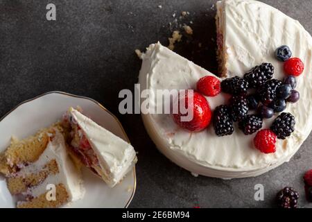 Immagine piatta di una torta rotonda con glassa bianca cremosa e assortimento di frutti di bosco utilizzati come guarnizioni decorative. Servita su sfondo nero grigio scuro. TW Foto Stock