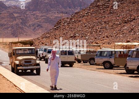 Un uomo beduino locale che indossa il keffiyeh e un abito da tobo arabo bianco di jubba cammina verso i veicoli safari fuoristrada nel deserto di Wadi Rum. Lui io Foto Stock