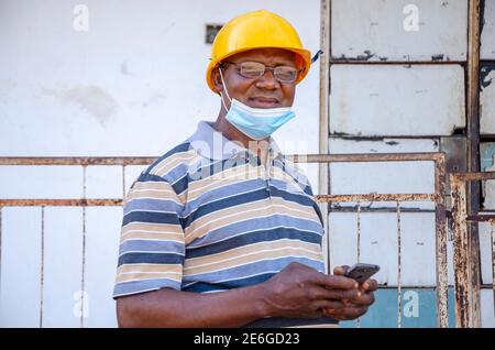 vecchio uomo nero che indossa una maschera al naso, indossa un casco e usa il suo smartphone Foto Stock