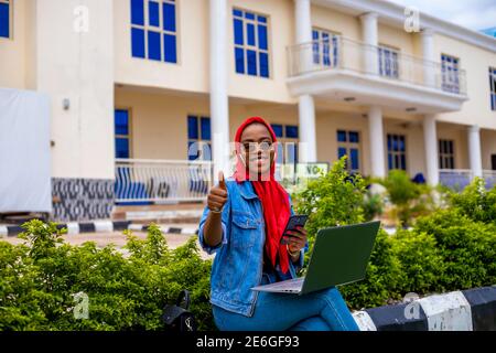 giovane donna nera bella seduta fuori e lavorando su di lei laptop e dare pollici in su Foto Stock