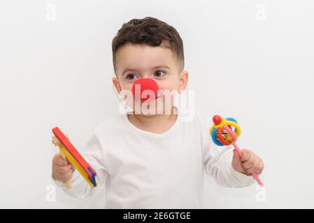 Felice capretto divertente che celebra Purim con un naso rosso e i noisemakers del legno. Ritratto di ragazzo ridente con un naso pagliaccio e gragnole su sfondo bianco. Focalizzazione morbida sui creatori di rumore per mostrare il movimento. Foto Stock