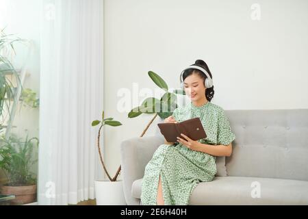 Giovane donna asiatica che ascolta musica dalle cuffie e nota di scrittura per la sua idea di lavoro nel diario libro. Foto Stock