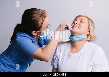Giovane infermiera in una maschera medica prende un test COVID 19 dal naso di una donna anziana su uno sfondo bianco. Foto Stock