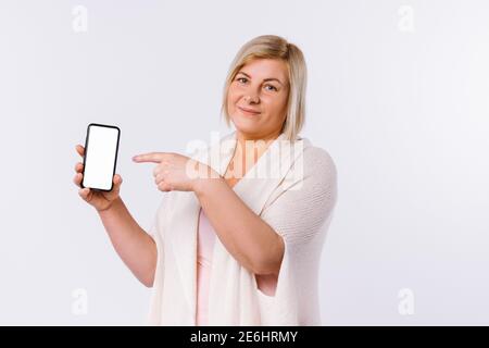 La donna bionda senior mostra il suo indice sullo schermo del telefono. Sfondo bianco e spazio laterale. Foto Stock