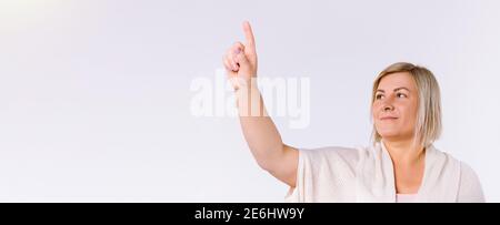 Banner, formato lungo. Una donna bionda mostra il dito indice verso l'alto e guarda di lato su uno sfondo bianco con spazio laterale vuoto. Foto Stock