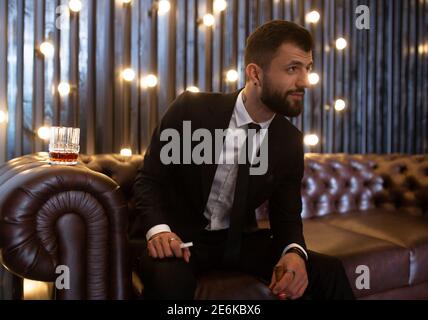 Ritratto di un uomo brutale barba con tatuaggio fumare sigaro in abito caldo elegante bere alcol whisky in appartamento loft . Concetto di storia d'amore di coppia Foto Stock