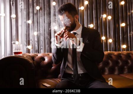 Ritratto di un uomo brutale barba con tatuaggio fumare sigaro in abito caldo elegante bere alcol whisky in appartamento loft . Concetto di storia d'amore di coppia Foto Stock