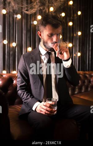 Ritratto di un uomo brutale barba con tatuaggio fumare sigaro in abito caldo elegante bere alcol whisky in appartamento loft . Concetto di storia d'amore di coppia Foto Stock