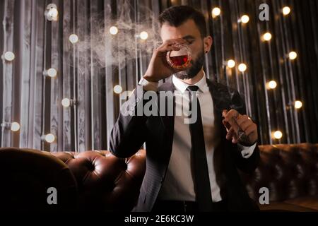 Ritratto di un uomo brutale barba con tatuaggio fumare sigaro in abito caldo elegante bere alcol whisky in appartamento loft . Concetto di storia d'amore di coppia Foto Stock