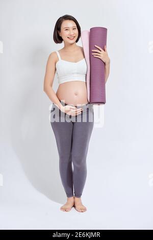 Giovane attraente donna incinta che tocca il suo ventre mentre tiene il tappeto di yoga nelle mani, gestante madre che fa esercizi fisici, posa isolata su W. Foto Stock