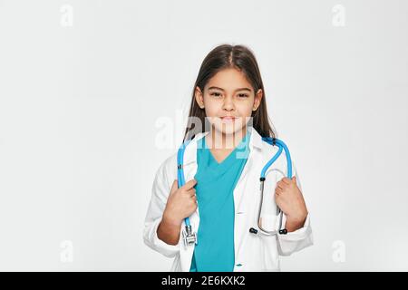 Ritratto di razza mista bambina vestita come medico con stetoscopio, lei posa e guardando la macchina fotografica. Sfondo grigio chiaro Foto Stock