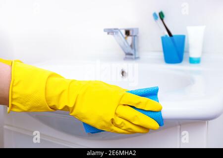 Ripulire la casa. Donna che fa i chores in bagno, mani in guanti gialli pulizia del rubinetto d'acqua, lavello in acciaio con straccio blu e spray detergente Foto Stock