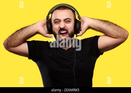 Urlante uomo arabico con barba ascolta la musica con le cuffie su sfondo giallo isolato. Giornata internazionale dei DJ Foto Stock
