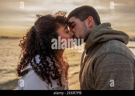 Testa e spalle ritratto di giovane bella coppia in amore baciando al tramonto in località balneare invernale con cielo nuvoloso. Due millenni in vacanza Foto Stock